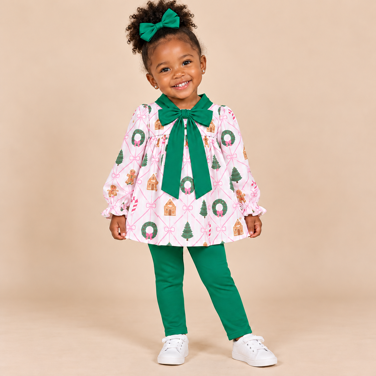 Toddler girl wearing a Christmas wreath and gingerbread house print top and green pants.