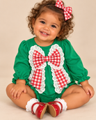 Baby wearing a green long-sleeve romper with a large red-and-white gingham bow, white eyelet trim, and ruffled cuffs.
