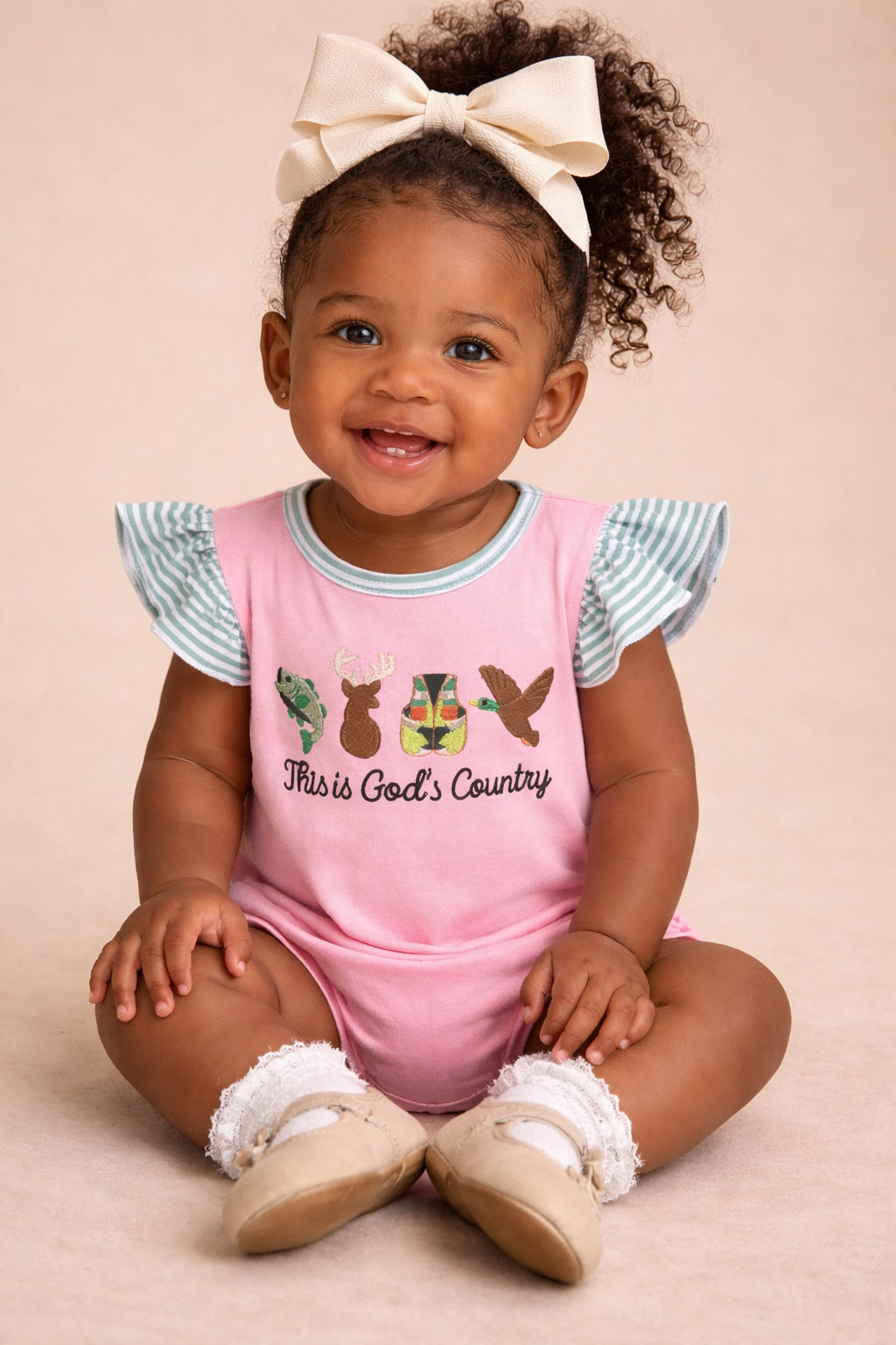 Child wearing a pink outfit with ruffled sleeves and 'This is God's Country' text, sitting on a beige background.