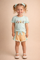 Young girl wearing a light blue top with cartoon characters and yellow checkered shorts on a beige background