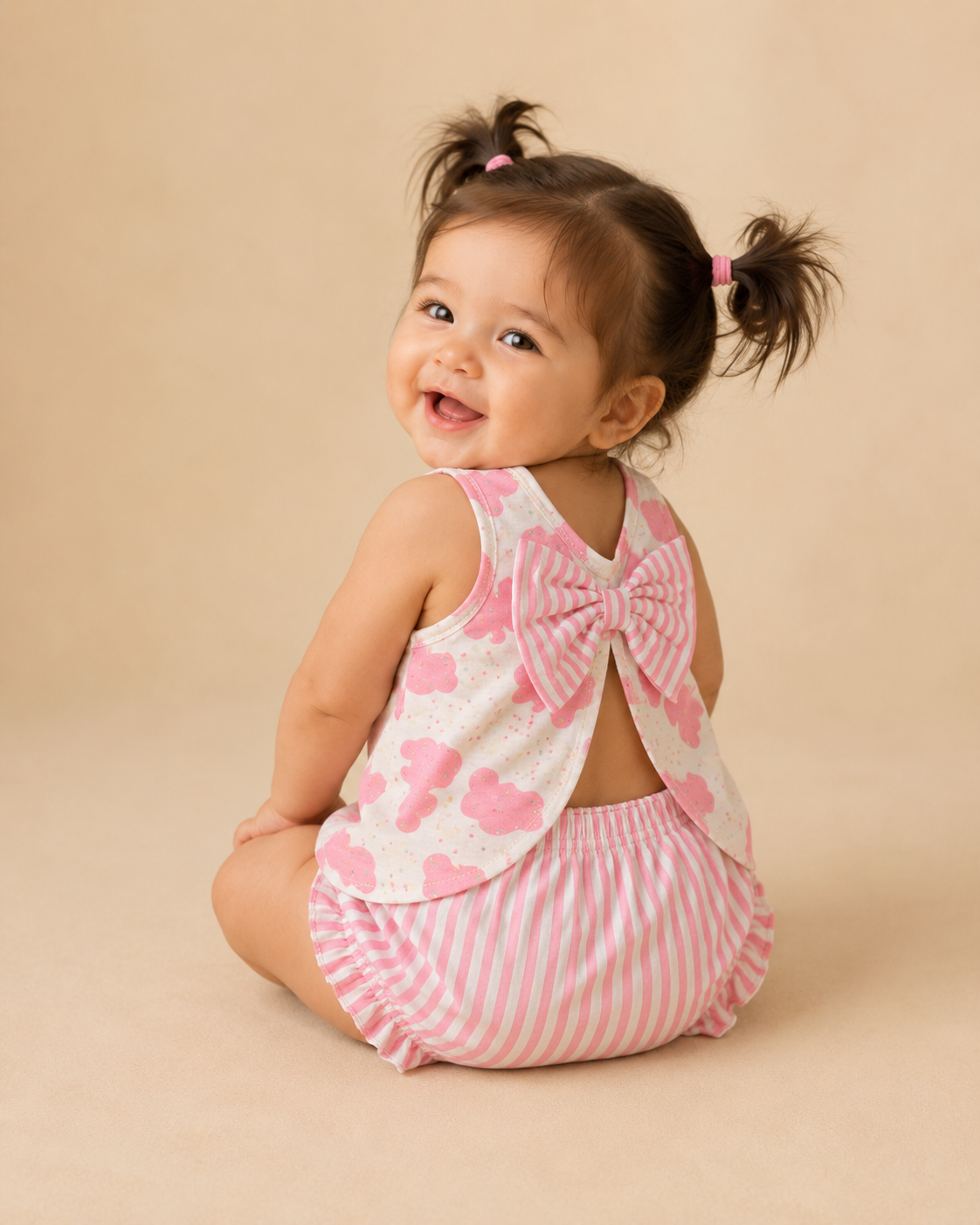 A baby girl sitting on a beige studio background, wearing a pink and white striped diaper set with a large bow on the back and a white top with pink animals.