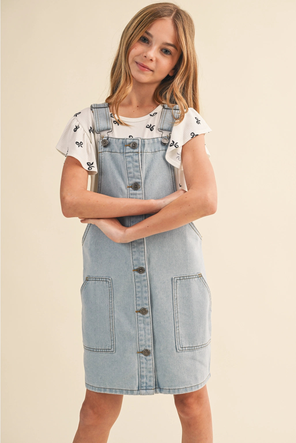 A young girl modeling a light blue denim overall dress over a white ruffle-sleeve t-shirt with black butterfly prints, set against a solid beige studio background.
