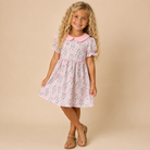 A young girl with long curly blonde hair stands against a solid beige studio background wearing a short-sleeve pink dress with a green and white floral pattern and a solid pink Peter Pan collar. She is wearing tan leather sandals.