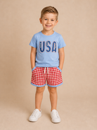 Child wearing a blue 'USA' t-shirt and red checkered shorts on a beige background