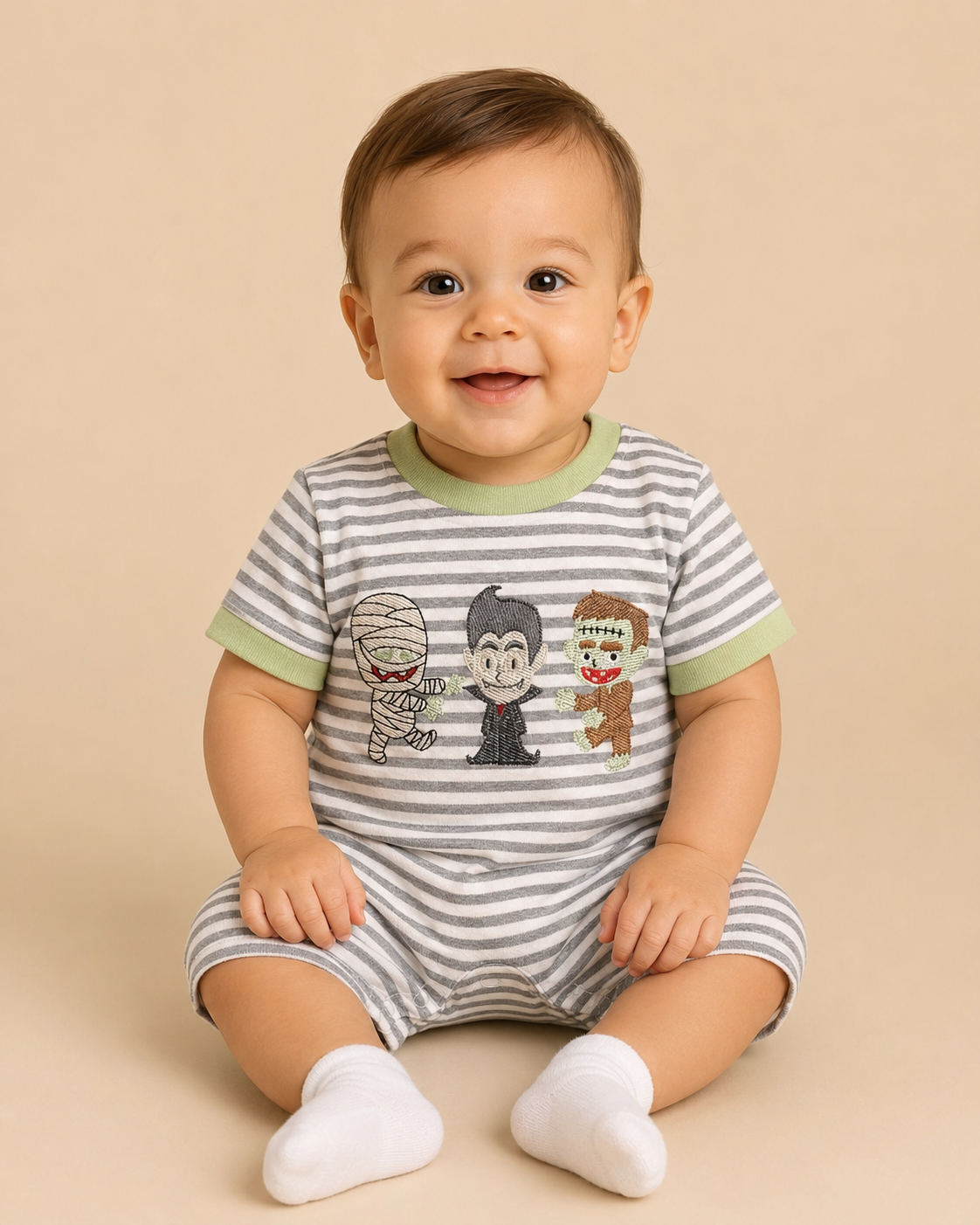 A smiling baby sitting on a beige studio background, wearing a gray and white striped short-sleeve romper with light green trim and a cartoon monster graphic on the front, along with white socks.