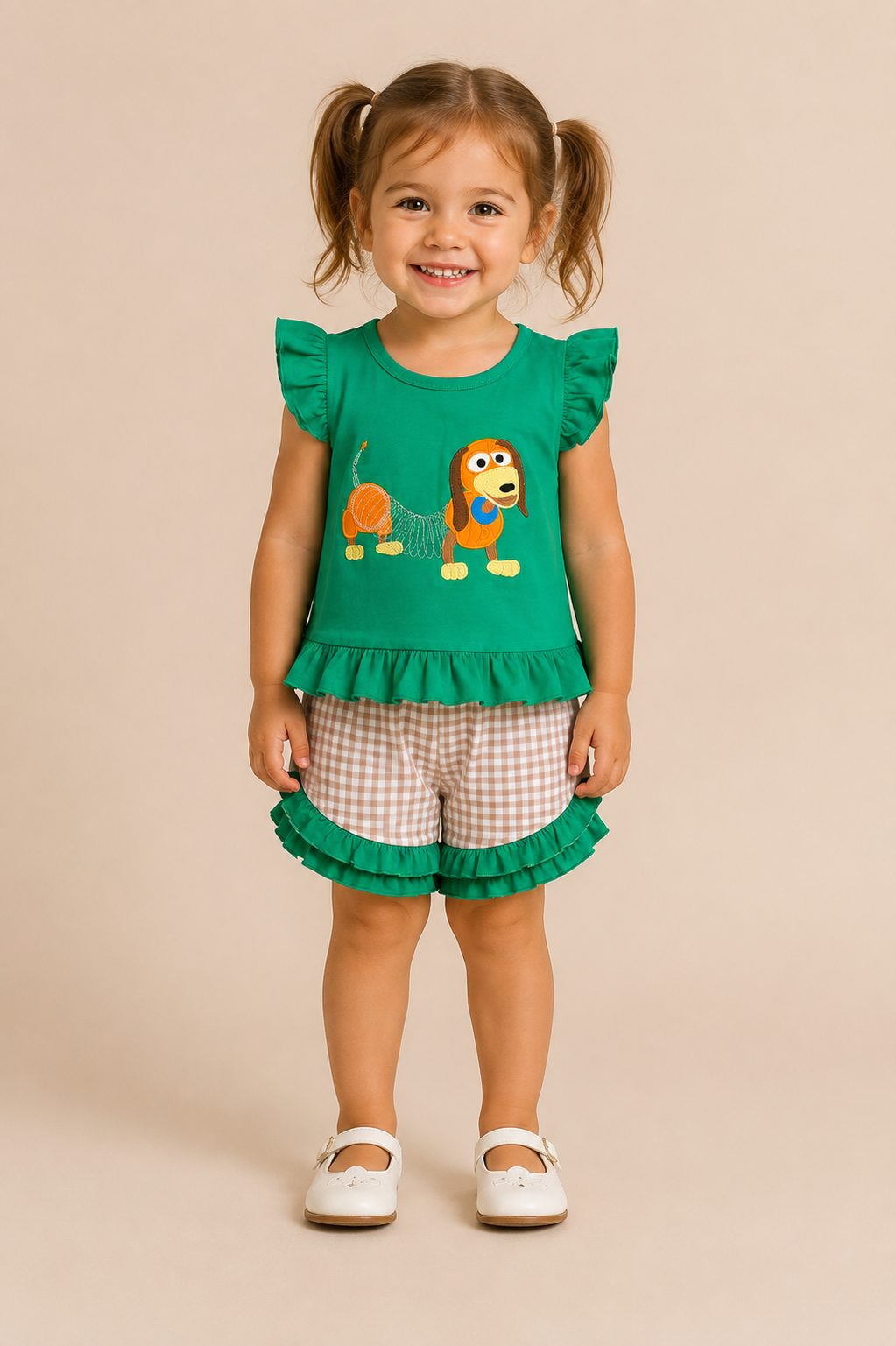 A young girl with pigtails stands against a solid beige background wearing a green ruffled sleeveless top with a cartoon dog graphic, paired with beige and white gingham shorts featuring a green ruffled hem, and white Mary Jane shoes.