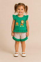 A young girl with pigtails stands against a solid beige background wearing a green ruffled sleeveless top with a cartoon dog graphic, paired with beige and white gingham shorts featuring a green ruffled hem, and white Mary Jane shoes.
