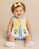 Baby wearing a blue checkered romper with yellow bows and a headband, sitting on a beige surface.