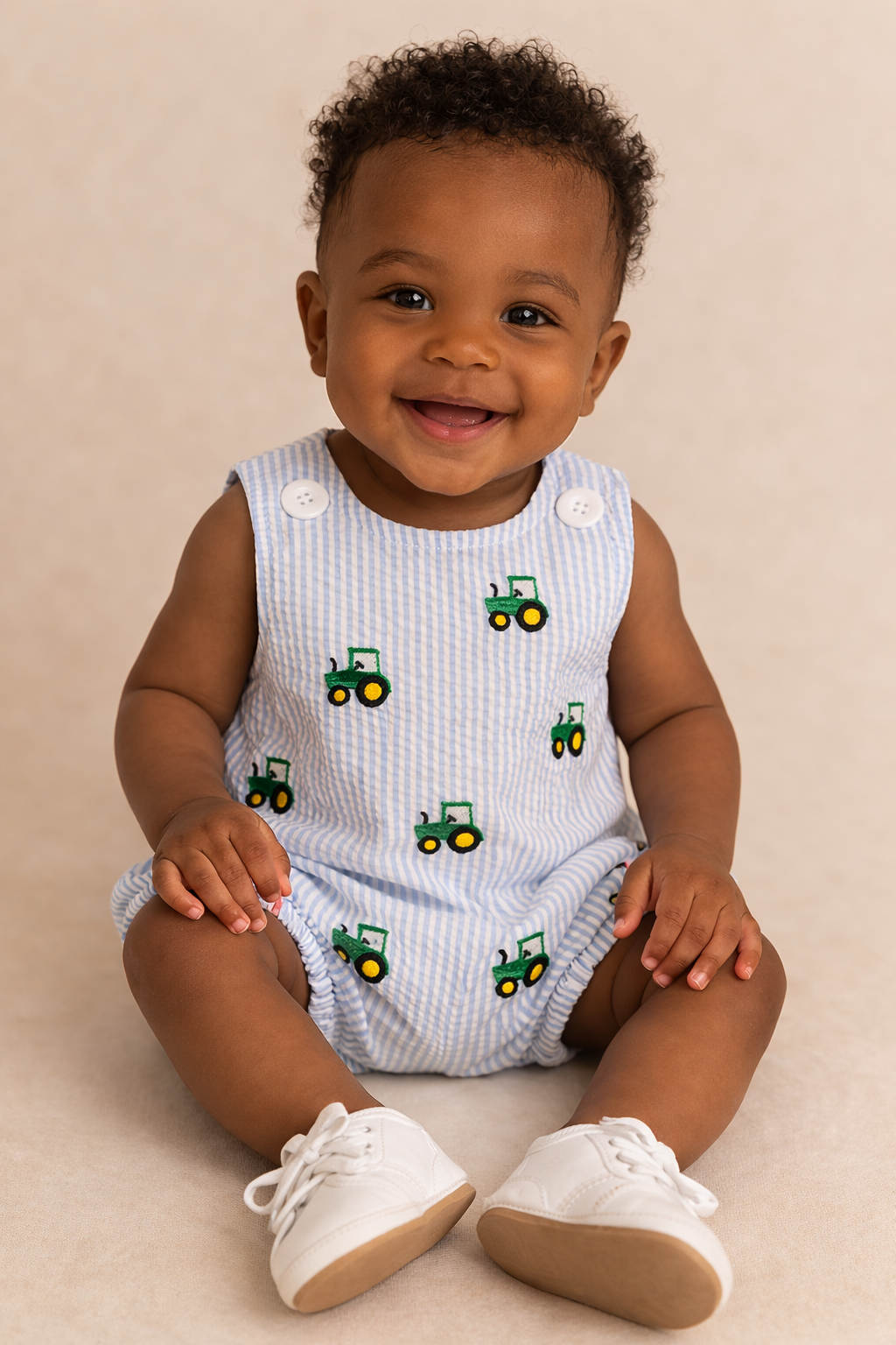 A baby boy sitting down wearing a light blue bubble with tractors on it.