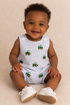 A baby boy sitting down wearing a light blue bubble with tractors on it.
