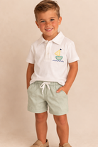 Child wearing a white polo shirt with a sailboat design and green checkered shorts on a beige background