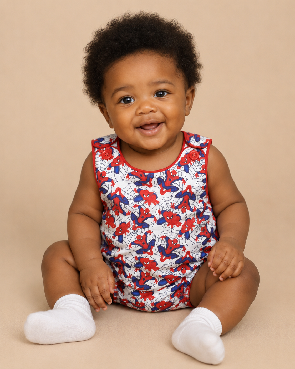A smiling baby sitting on a beige studio background, wearing a sleeveless white romper covered in a red and blue spiderweb pattern, with white socks.