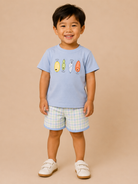 A young child wearing a light blue short-sleeve t-shirt with a colorful fish graphic and light blue and white gingham shorts, standing against a solid beige background.