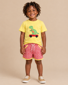 Toddler boy wearing a yellow shirt with a dinosaur on the front and red gingham shorts with yellow trim.