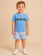 A young boy stands against a solid beige studio background wearing a light blue short-sleeve t-shirt with a green tractor graphic, matching blue and white striped shorts, and white sneakers.
