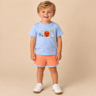 Toddler boy wearing a Halloween themed shirt, light blue with orange gingham shorts on a beige background.