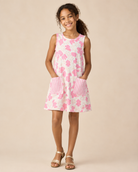 A young girl stands against a solid beige studio background wearing a sleeveless white dress with a pink floral pattern, pink-and-white striped pockets, and tan sandals.
