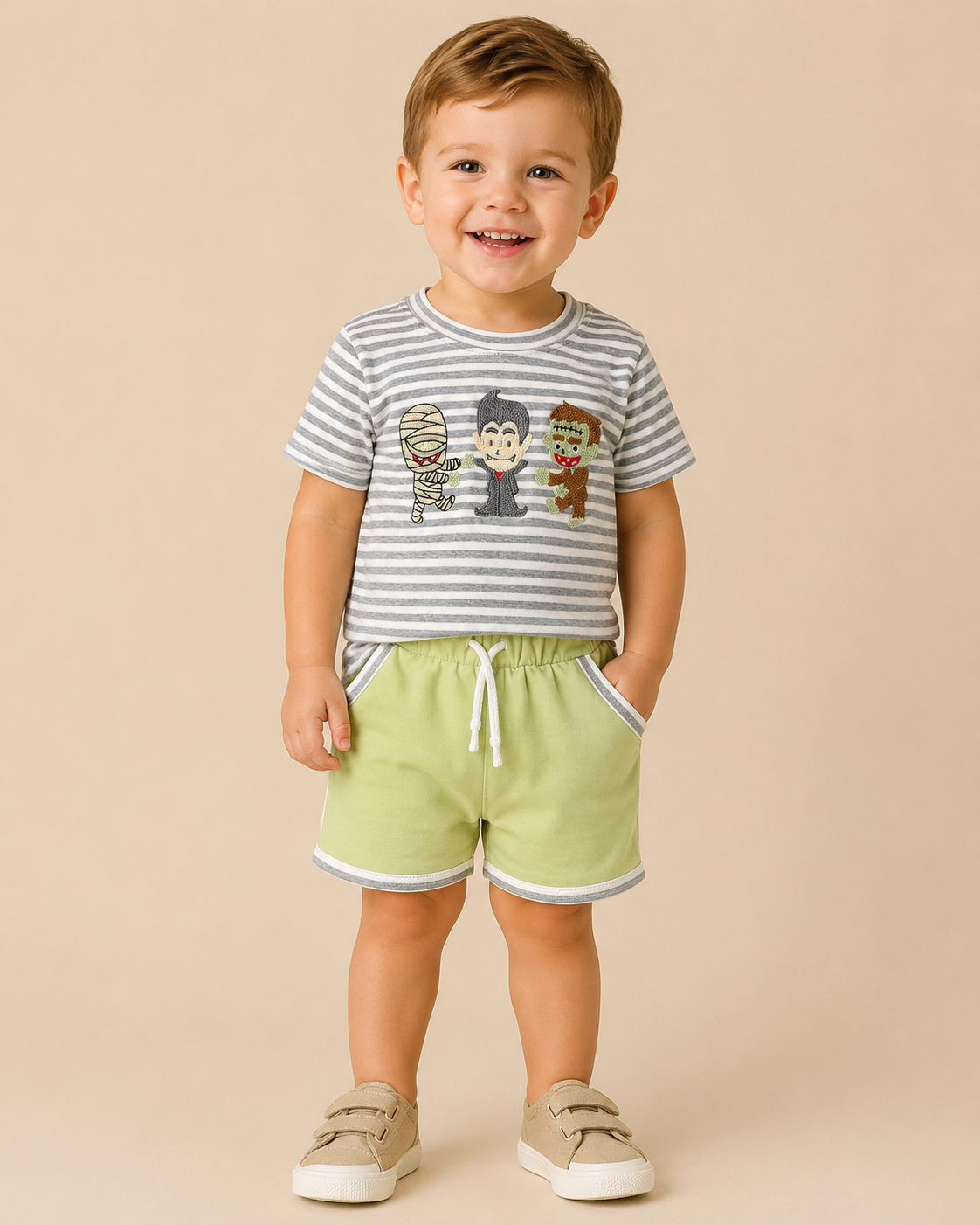A smiling toddler standing against a plain beige studio background, wearing a gray-and-white striped short-sleeve T-shirt with cartoon character graphics on the chest, light green shorts with white trim, and beige slip-on shoes.