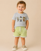 A smiling toddler standing against a plain beige studio background, wearing a gray-and-white striped short-sleeve T-shirt with cartoon character graphics on the chest, light green shorts with white trim, and beige slip-on shoes.