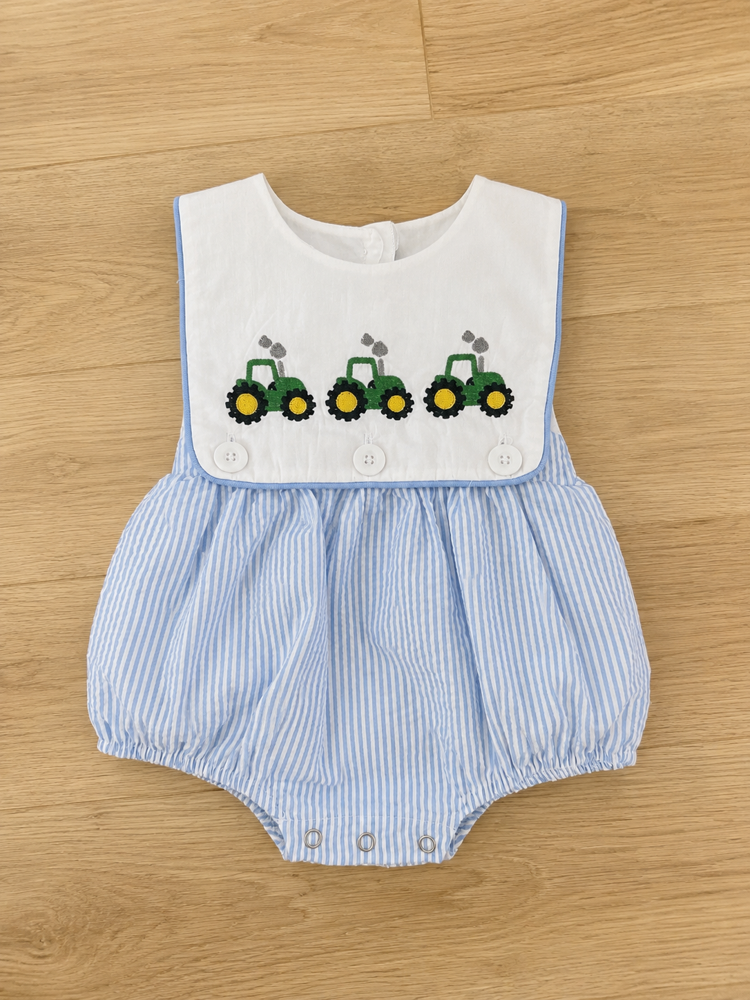 A baby bubble romper laid flat on a light wood floor. The romper has a white bib-style top with three embroidered green tractors with yellow wheels and gray smokestacks, and a light blue and white vertically striped bodice and leg sections.