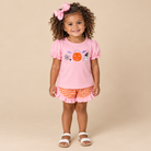Toddler girl wearing a pink shirt with Halloween themed embroidery. Orange gingham shorts on a beige background.
