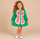 Girl wearing a green long-sleeve Christmas dress with an oversized red-and-white gingham bow, white eyelet trim, and ruffled cuffs.