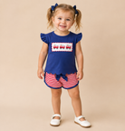 Toddler girl with pigtails wearing a blue flutter sleeve shirt with firetrucks embroidered and red & white shorts. 