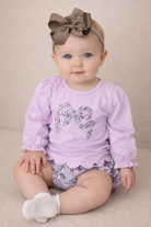Baby girl sitting down, wearing a lilac top with a floral bow in the middle.