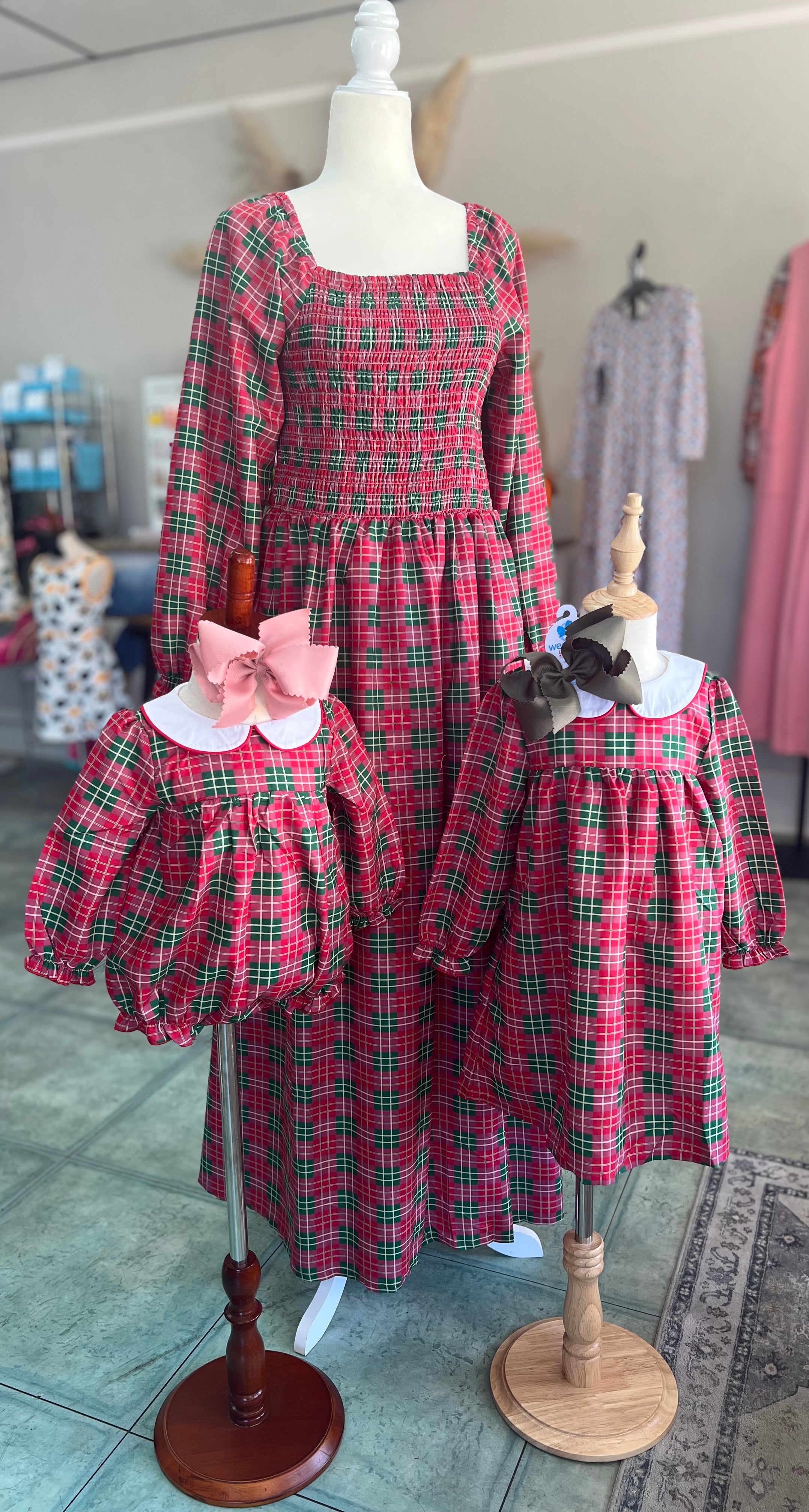 A festive matching family set displayed on mannequins inside a boutique. The set features a red and green plaid pattern, including a long-sleeve smocked women’s dress with a square neckline, a girls’ long-sleeve dress with a white Peter Pan collar and brown bow, and a coordinating baby bubble romper with the same plaid fabric, white collar. Perfect for family photos or holiday events.