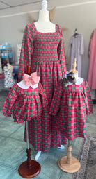 A festive matching family set displayed on mannequins inside a boutique. The set features a red and green plaid pattern, including a long-sleeve smocked women’s dress with a square neckline, a girls’ long-sleeve dress with a white Peter Pan collar and brown bow, and a coordinating baby bubble romper with the same plaid fabric, white collar. Perfect for family photos or holiday events.