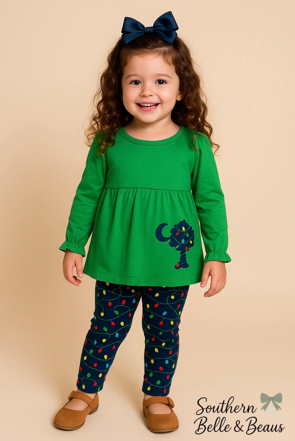 Little girl wearing Palmetto Christmas outfit with green top and Christmas light leggings, navy bow, and tan shoes on beige background.