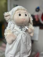 Plush lamb toy dressed in a white outfit with a cross, against a blurred background.