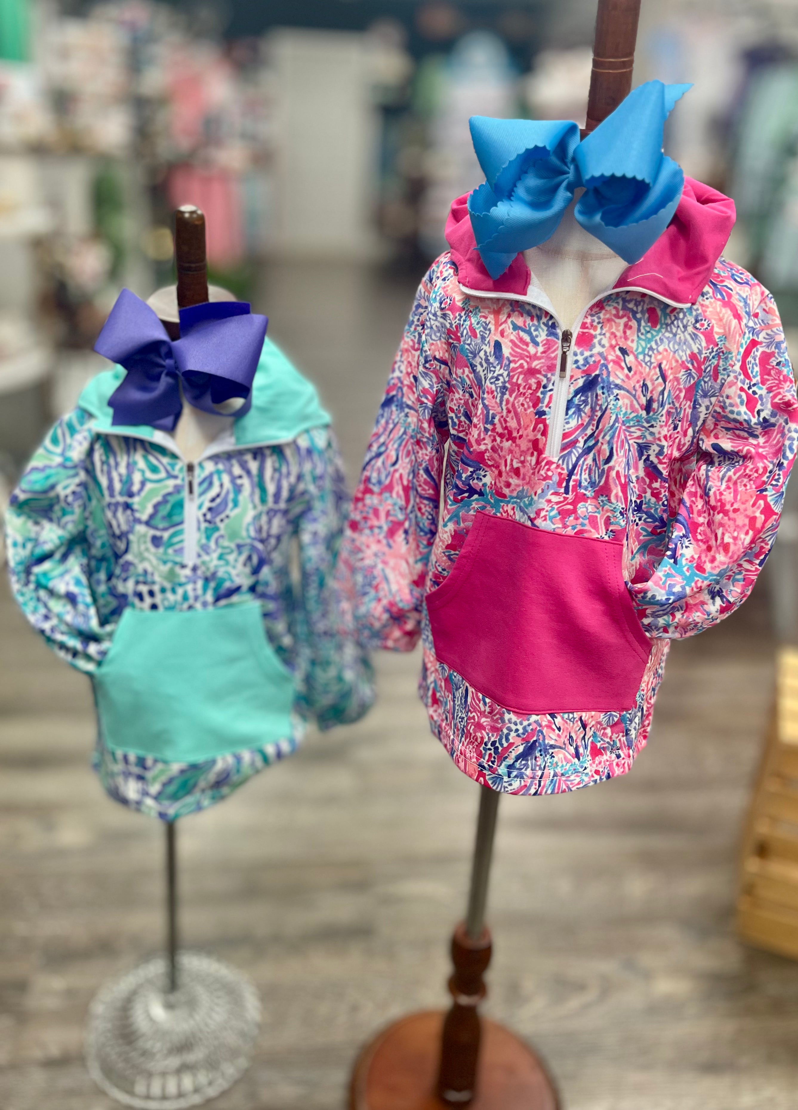 Two children's jackets with large bows on mannequins against a blurred indoor background.