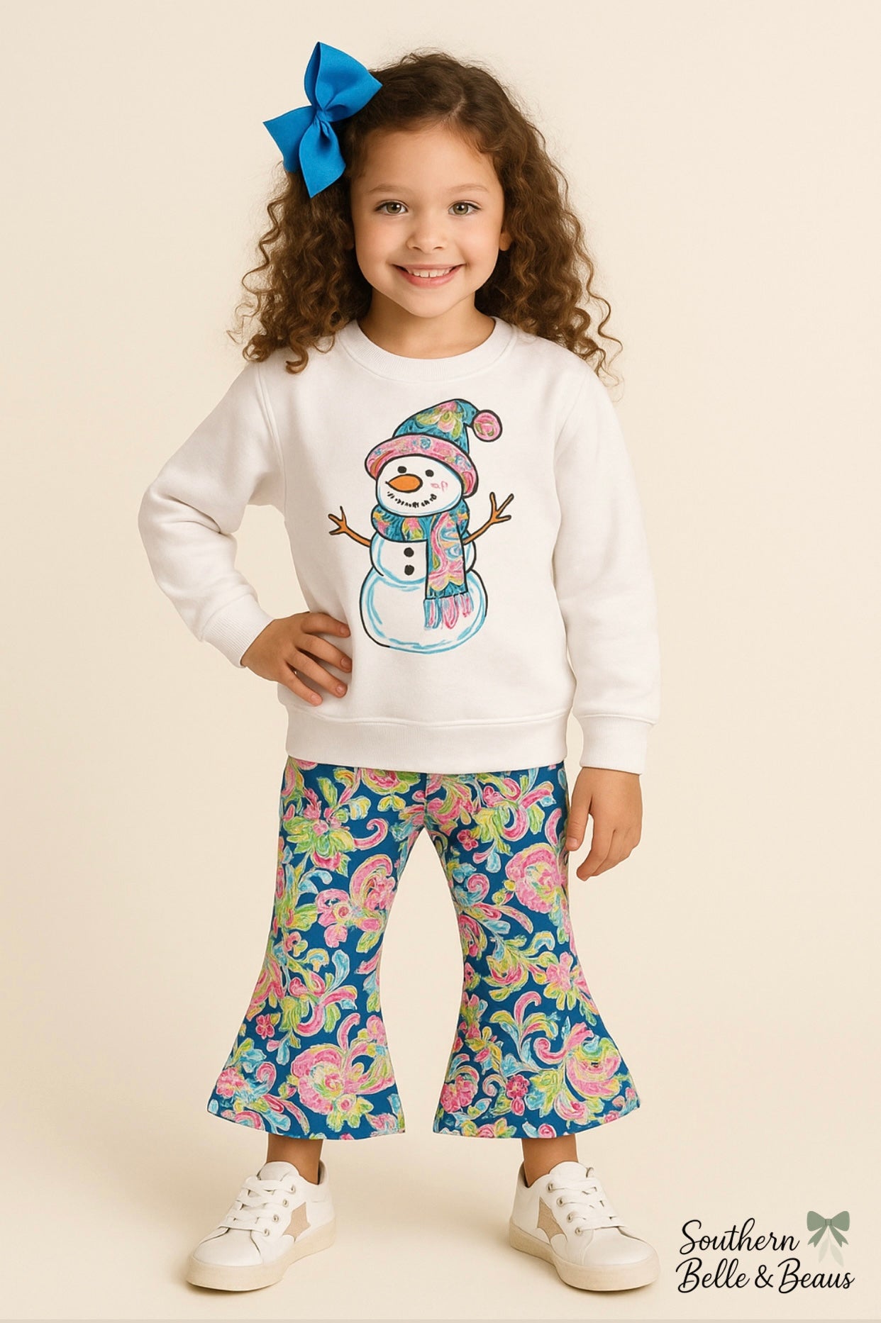 Young girl wearing a white sweatshirt with a snowman design and colorful floral pants on a beige background.