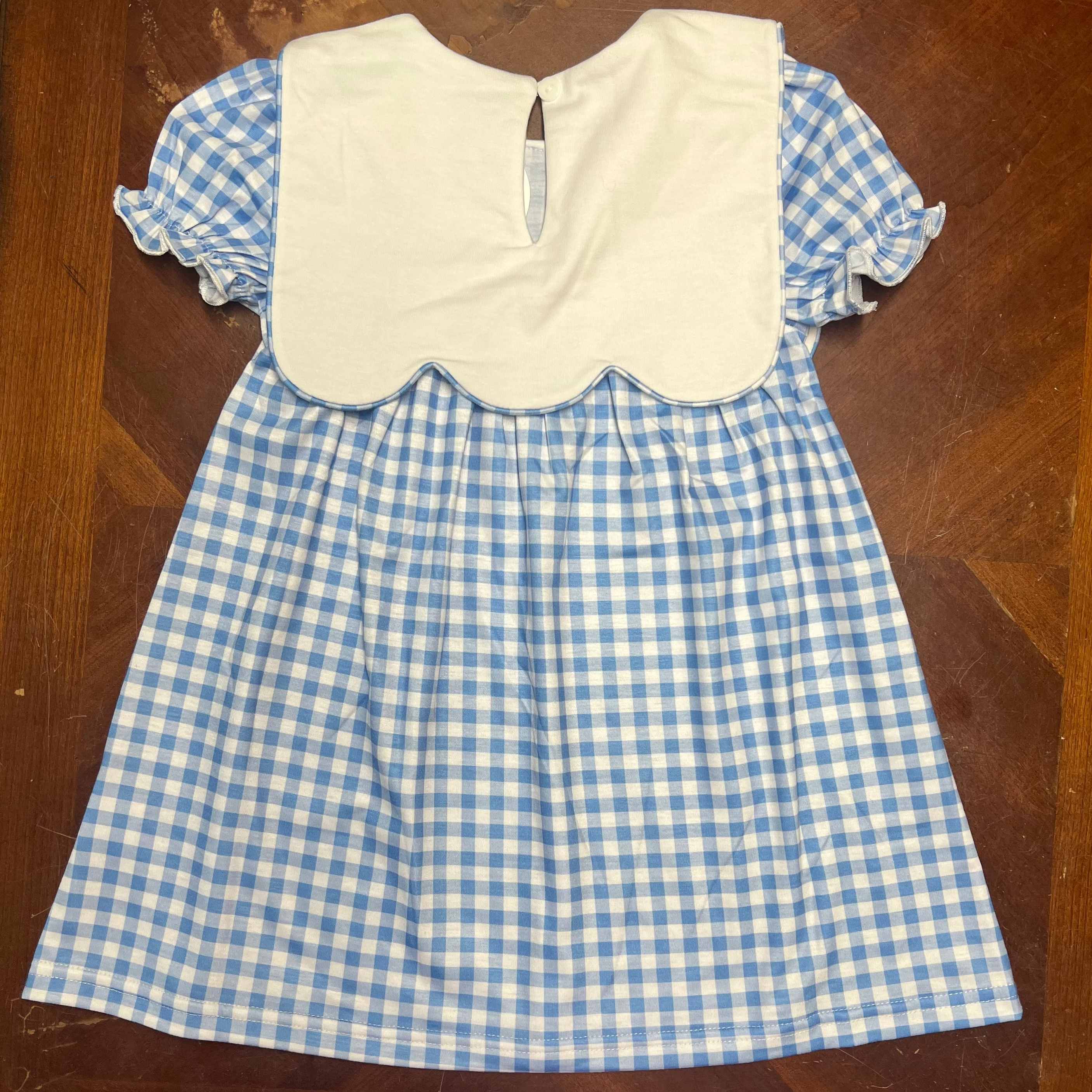 A children's dress laid flat on a wooden table, featuring a white yoke with a scalloped edge and short blue-and-white gingham sleeves and skirt.