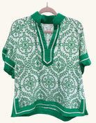 Green and white geometric patterned top with split neckline, contrast trim, and short sleeves, featuring a relaxed fit and lightweight fabric.