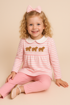 Young girl wearing a pink and white striped tunic top with embroidered dogs and pink pants, sitting on a beige background.