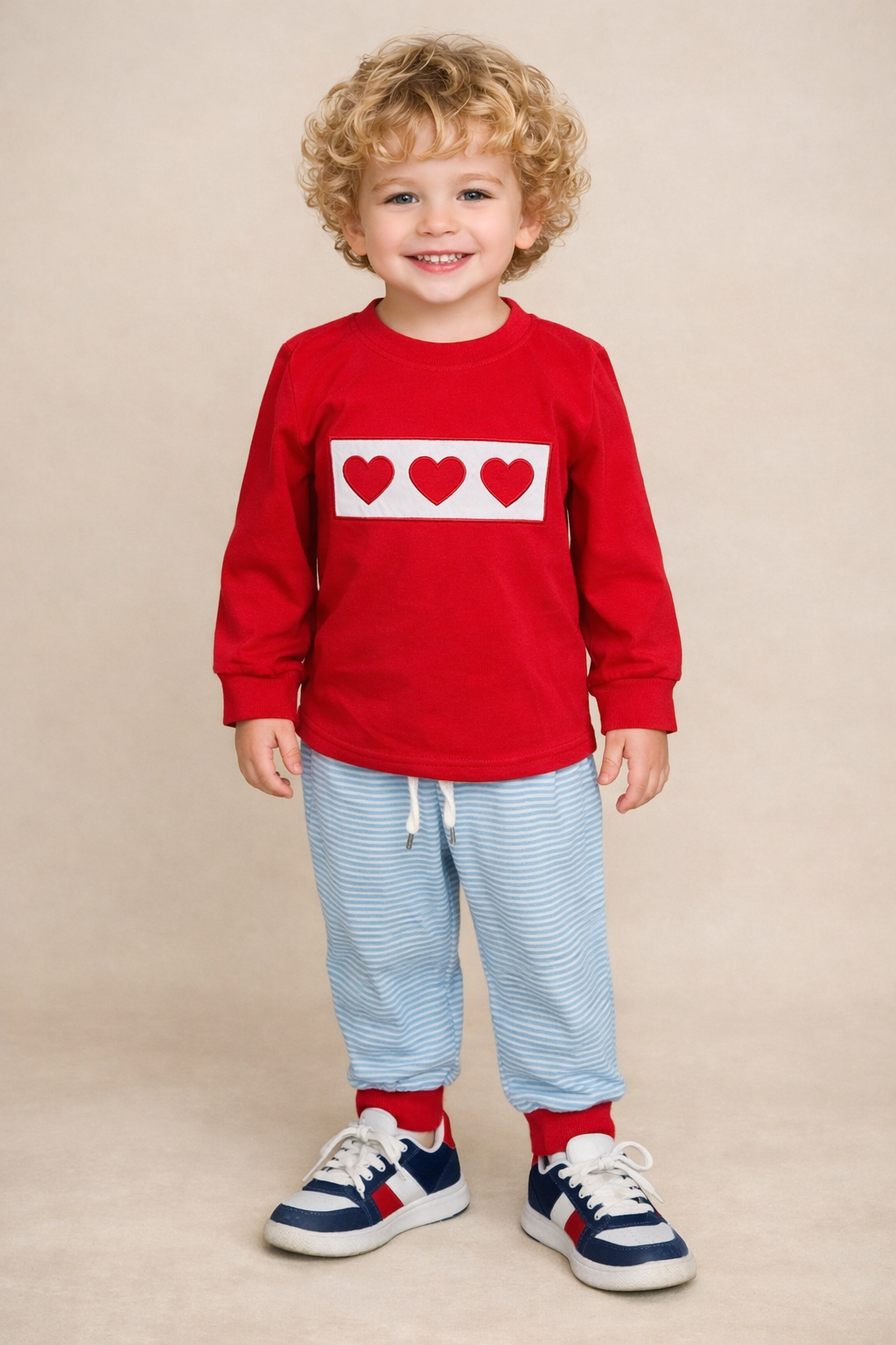 Boy wearing a red sweatshirt with heart designs and light blue pants on a beige background.