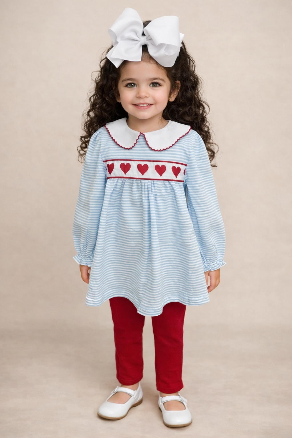 Young girl wearing a blue and white striped dress with red heart details and red pants on a beige background