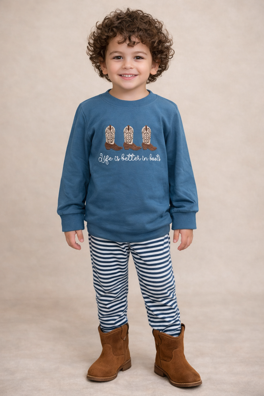 Curly headed boy wearing a long sleeve shirt with boots on the front and blue and white striped joggers. 