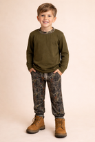 A boy wearing a dark green shirt with camo joggers and boots.