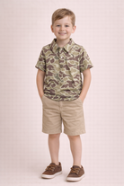 Young boy wearing a camouflage shirt and beige shorts on a light pink background