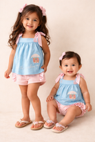 Two young girls wearing matching blue tops and pink shorts with ruffles on a plain background.