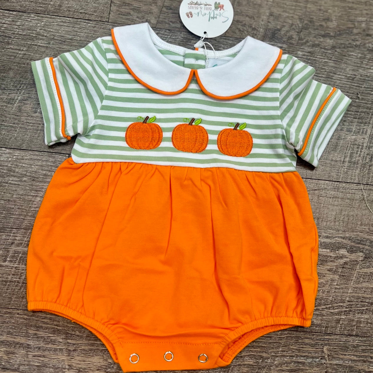 A baby short-sleeve romper laid flat on a dark wood floor. The top half has light green and white horizontal stripes with three orange pumpkin appliques across the chest and a white Peter Pan collar outlined in orange.