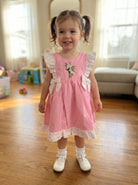 Young girl in a pink dress standing on a wooden floor in a home setting.