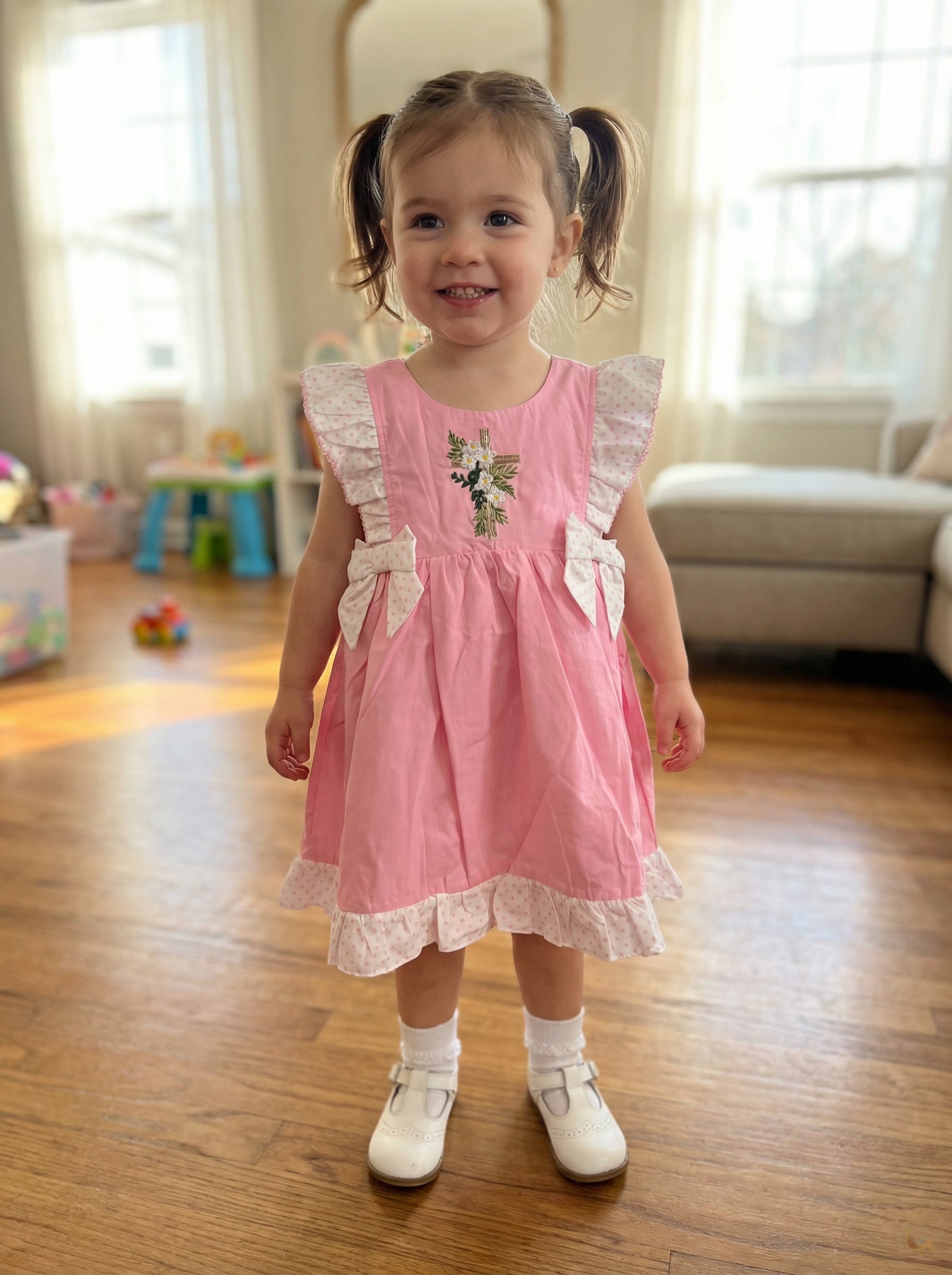 Young girl in a pink dress standing on a wooden floor in a home setting.