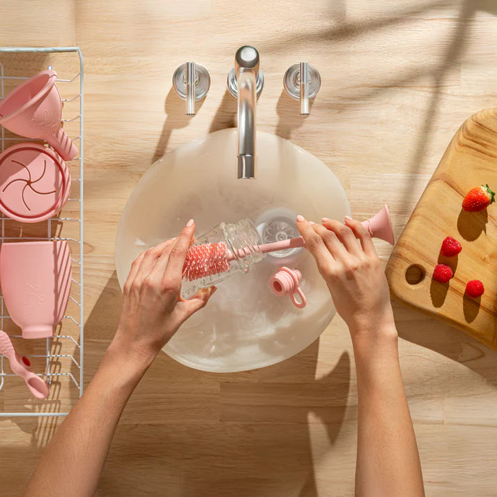 Person cleaning a bottle with a brush in a sink, with pink kitchen items and a wooden cutting board in the background.
