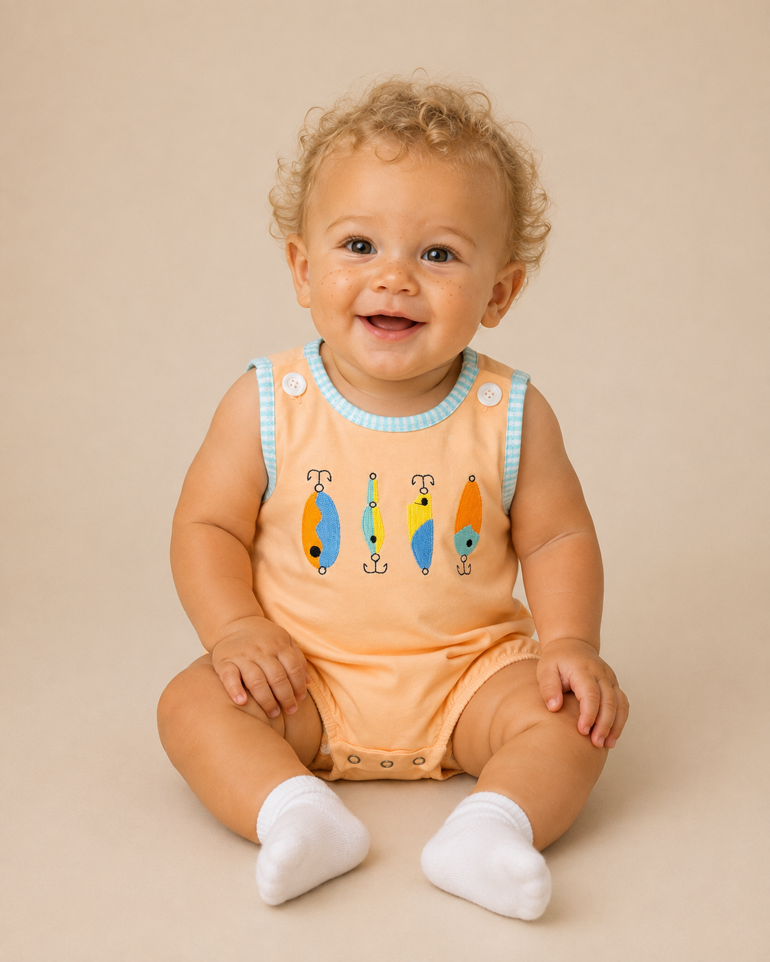 A smiling baby with curly blonde hair sits on a beige studio background wearing a peach sleeveless bubble with light blue trim and colorful fish embroidery, along with white socks.