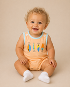 A smiling baby with curly blonde hair sits on a beige studio background wearing a peach sleeveless bubble with light blue trim and colorful fish embroidery, along with white socks.