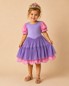 A young girl wearing a purple and pink princess costume dress with a gold tiara and sandals, standing against a solid beige studio background.